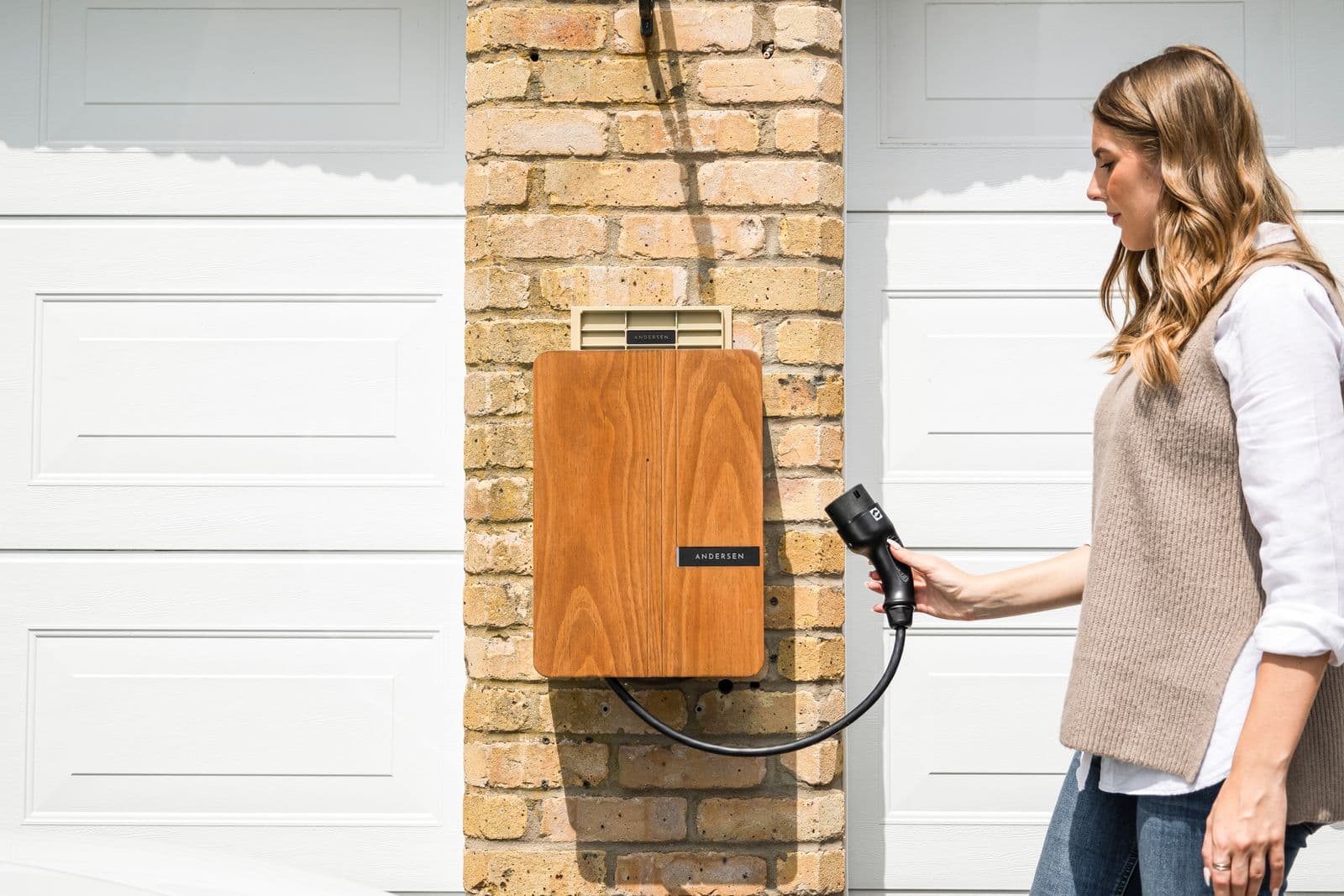 Person holding a connector beside a wall-mounted home EV charger on a brick wall