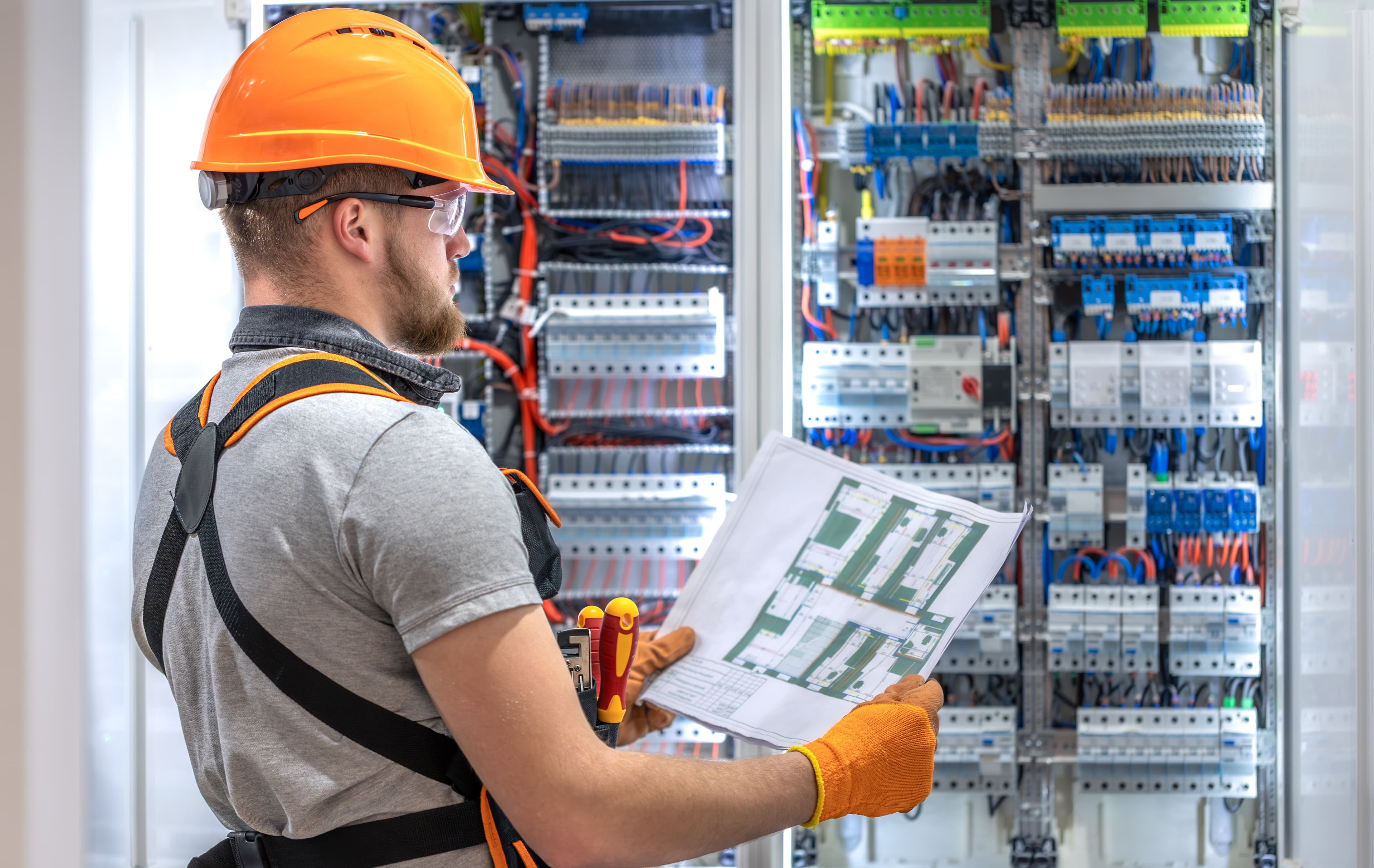 Licensed electrician in safety gear reviewing a wiring schematic in front of an electrical distribution panel